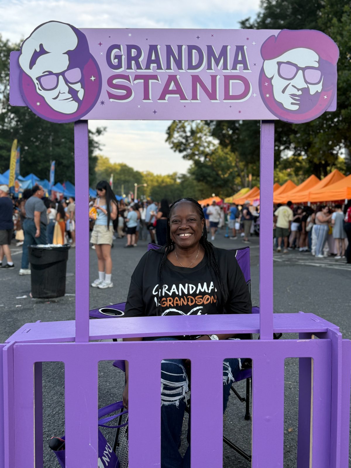 A smiling grandma sits behind the purple stand at a street fair